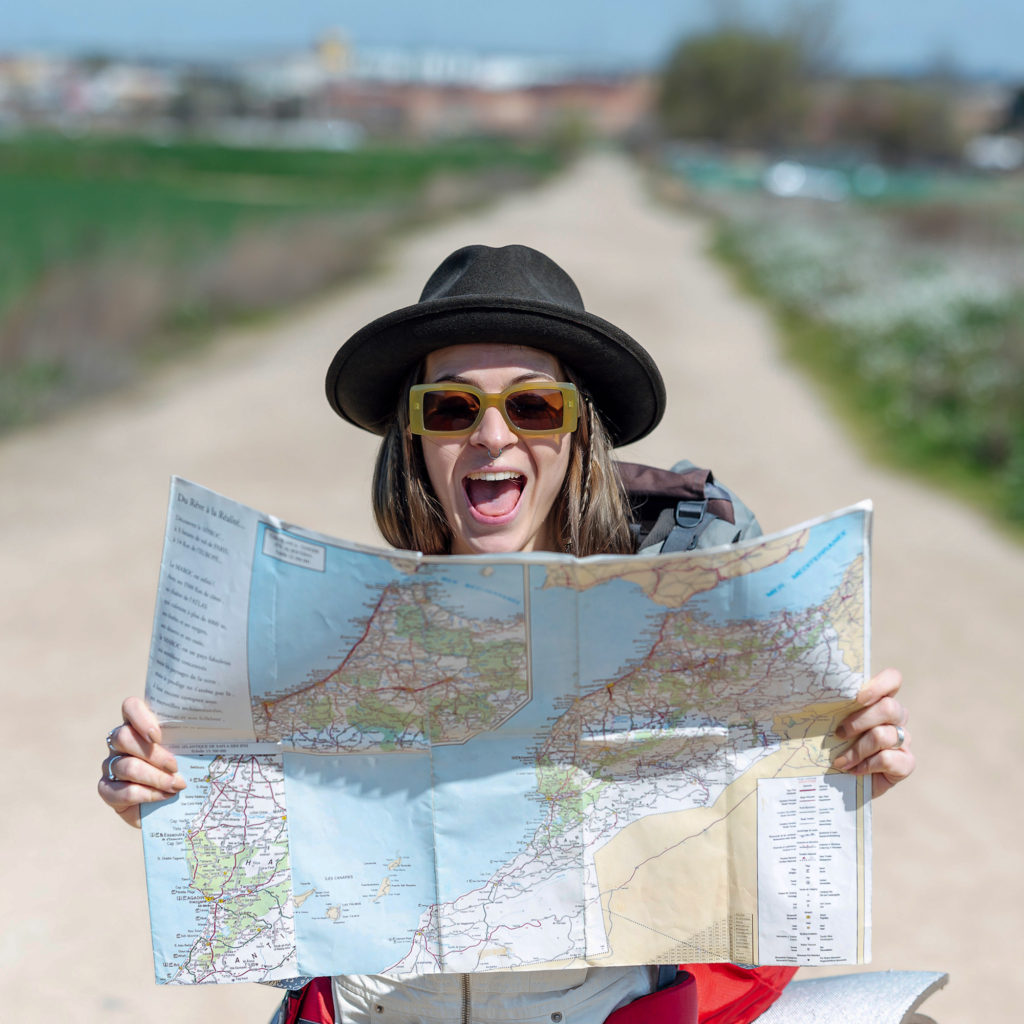 Young woman with yellow sunglasses and dark hat wearing a backpack looks at map in her hands